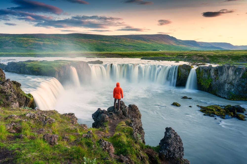 Godafoss waterfall flowing with sunset sky and male tourist in a orange coat
